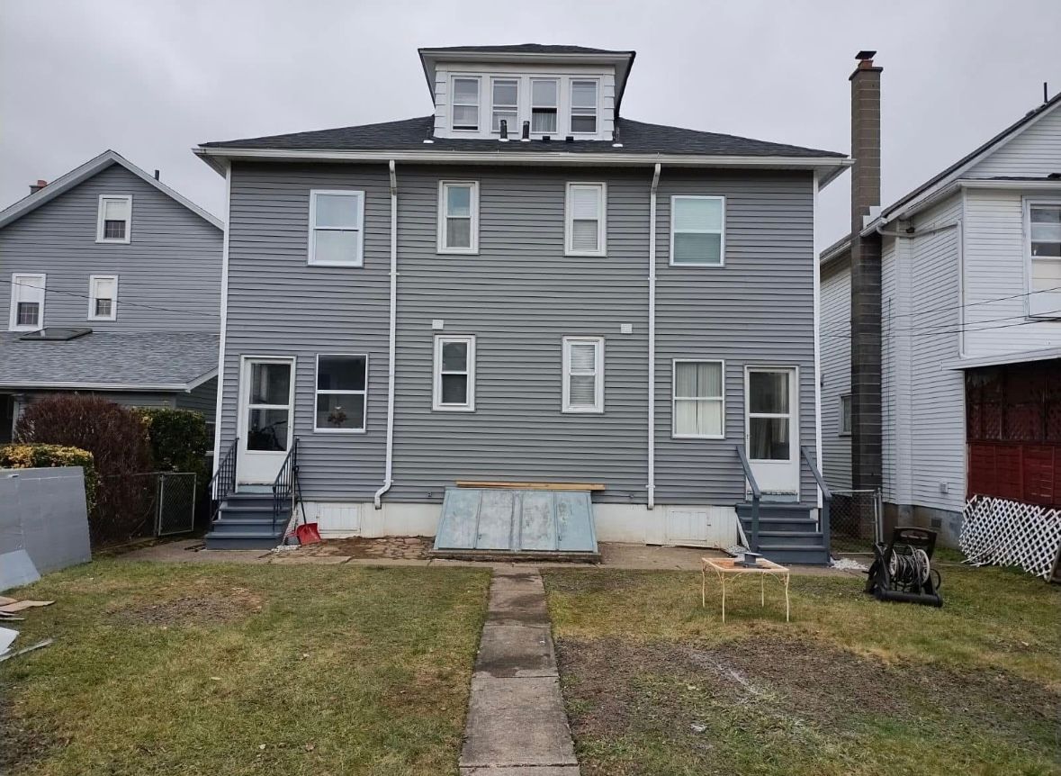 Gray two-story house with white trim, front lawn, and concrete walkway, seen from the backyard.