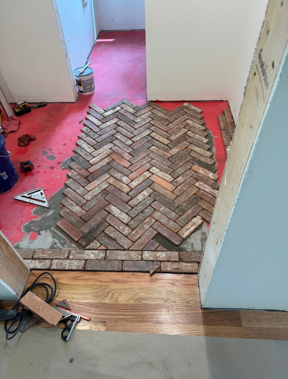 Hallway under renovation with a herringbone brick floor section and tools on the red underlayment.