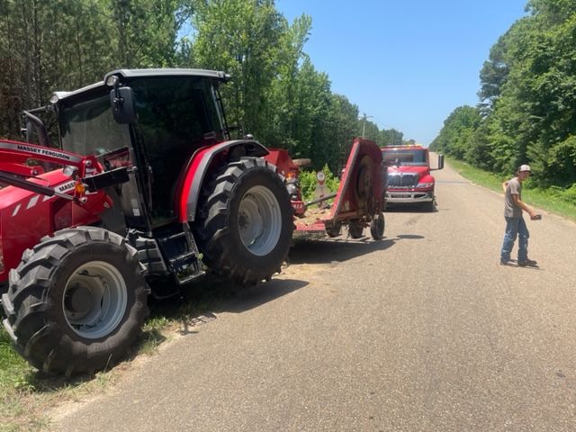 Red tractor with mower on a road, a semi-truck behind it, and a person standing nearby.