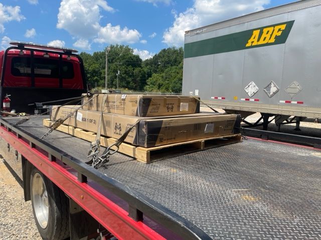 Tow truck with a pallet of boxes secured to its bed, beside a semi-truck with ABF logo.