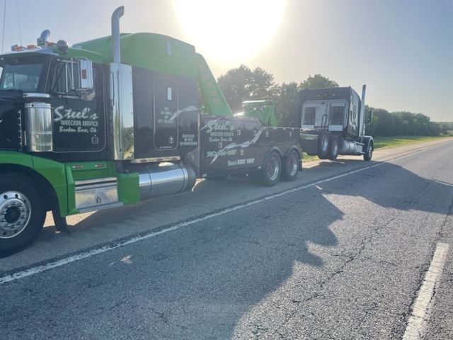 A green and black tow truck towing a semi-truck on a highway, sunny day.