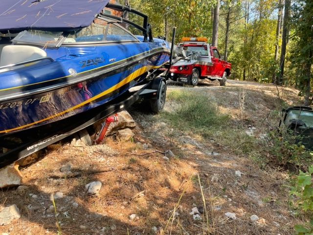 Blue boat on trailer being towed by a red tow truck on a dirt road.