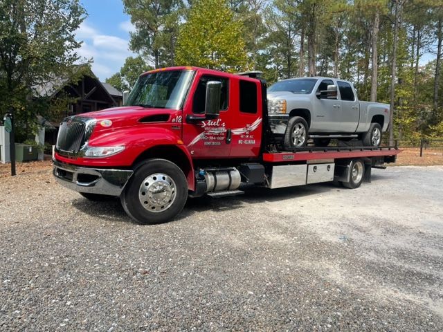 Red tow truck transporting a silver pickup truck on a gravel driveway.