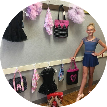 A dancer in a blue leotard and skirt stands next to a wall with racks displaying dance clothes, bags, and shoes.