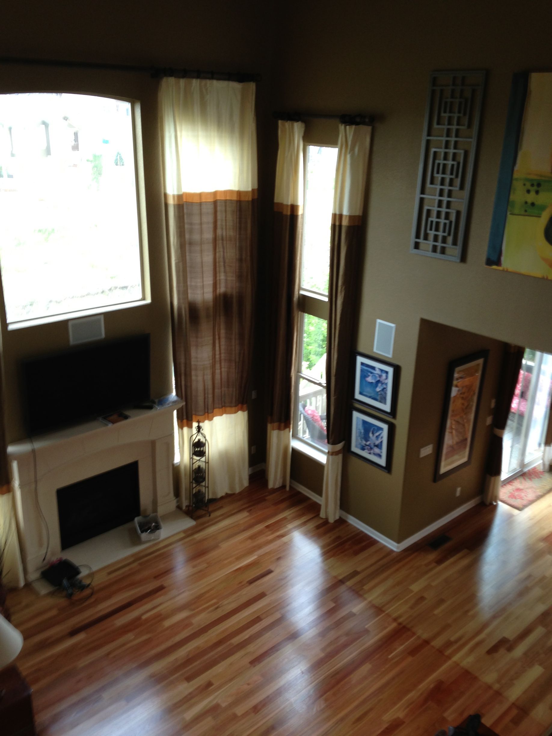 High-angle view of a living room with tall windows, brown curtains, fireplace, wooden floors, and neutral-colored walls.