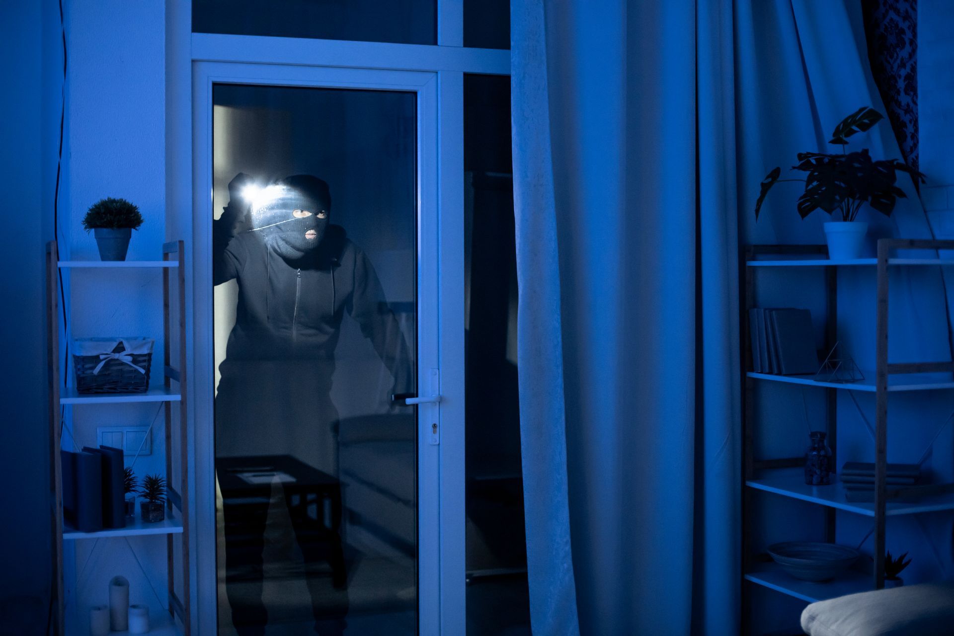Burglar wearing a mask and holding a flashlight is visible through a glass door at night.