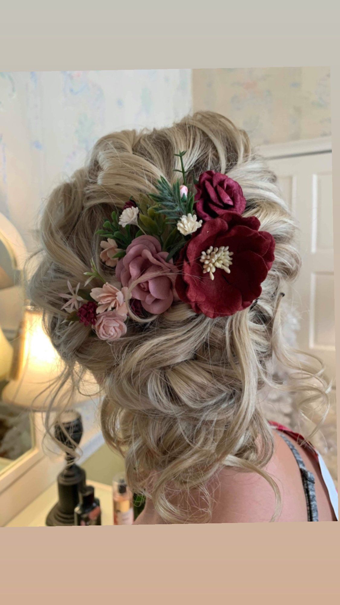 Woman's blonde updo hairstyle with burgundy and pink flowers. Indoors, vanity in background.