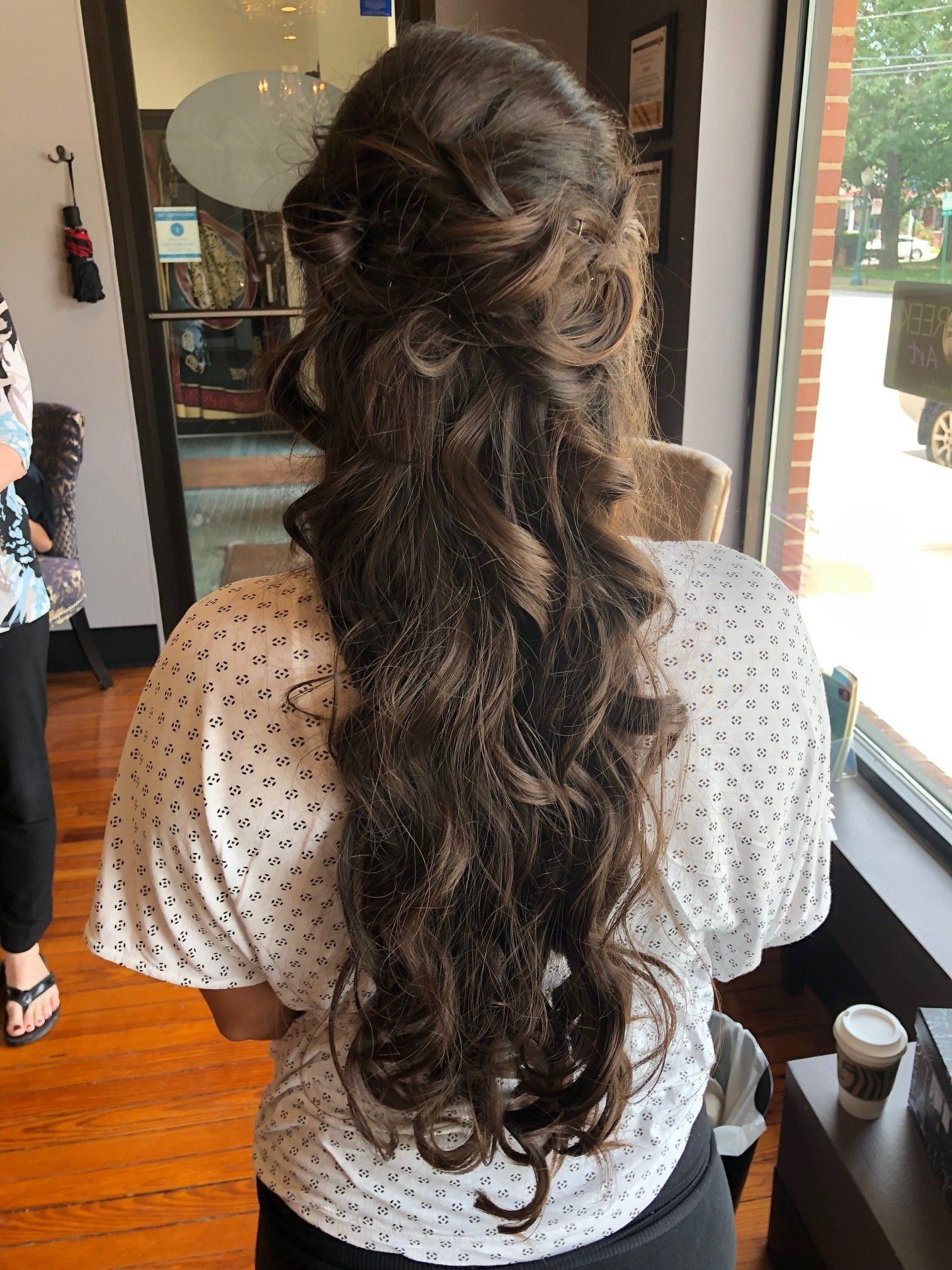 Woman with long brown wavy hair styled in a half-updo, standing in front of a window.