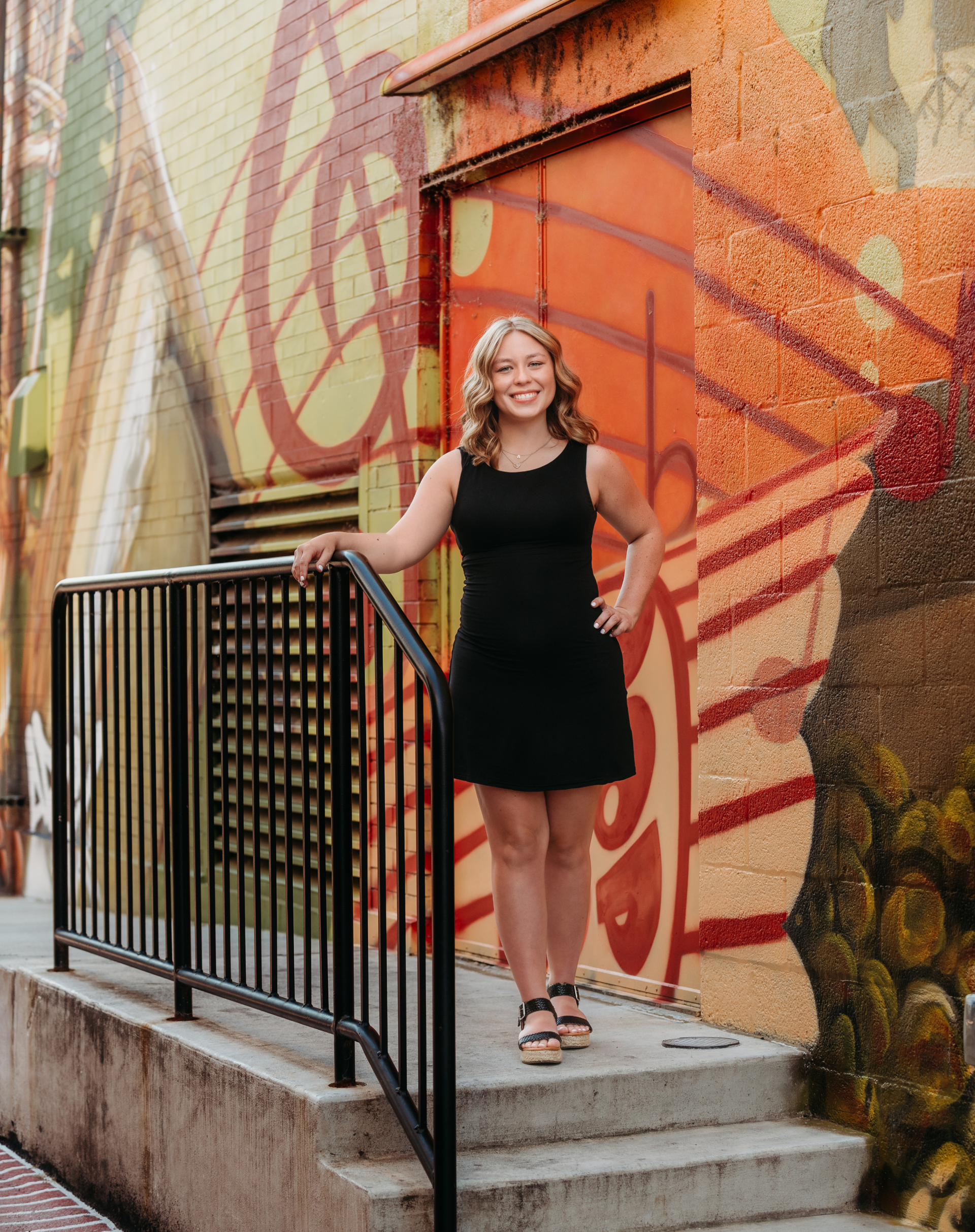 Young woman with red hair smiles, leaning on a bridge railing, wearing a black dress. River in background.