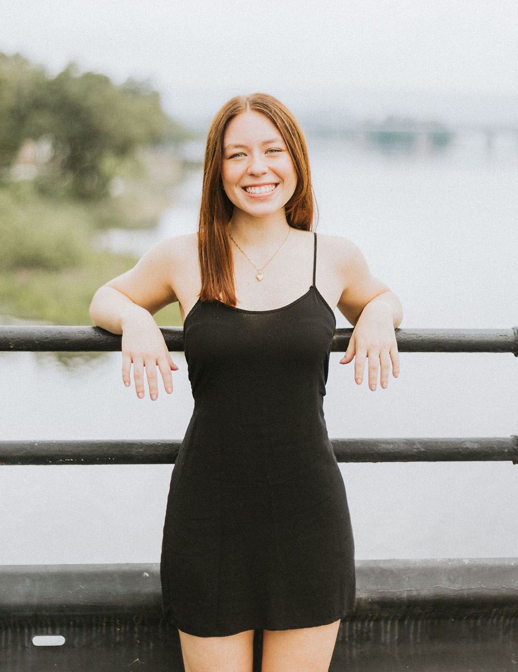Young woman with red hair smiles, leaning on a bridge railing, wearing a black dress. River in background.