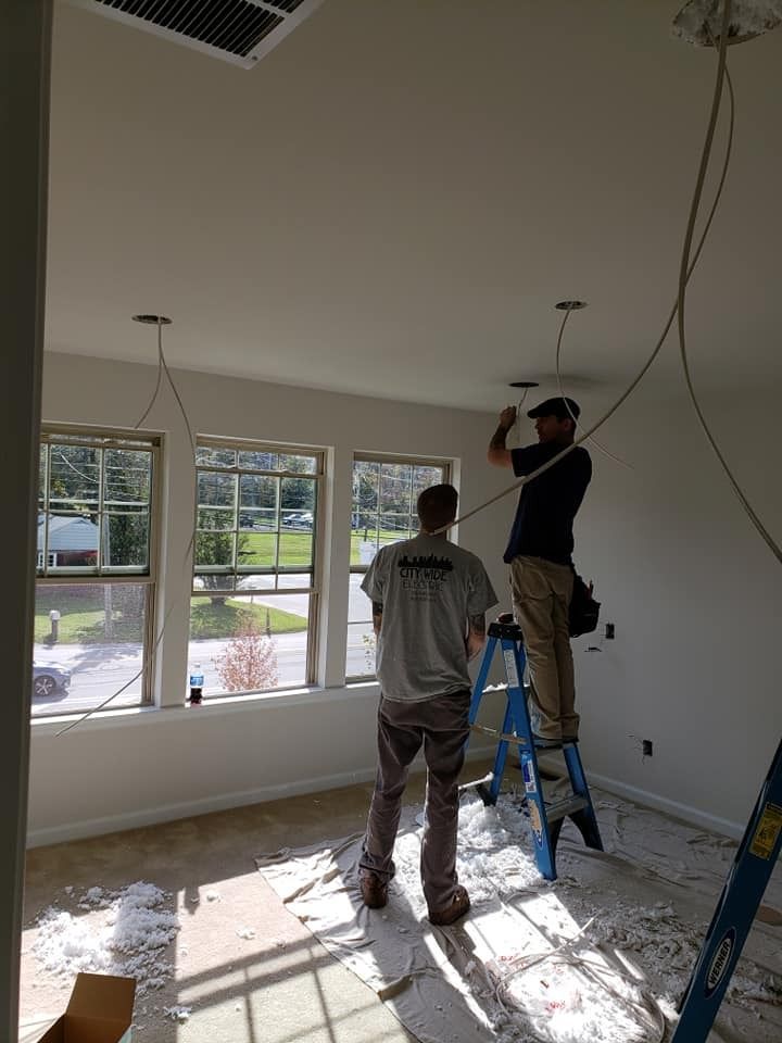 Two workers installing ceiling lights in a room with windows. One stands on a ladder, wires hanging.