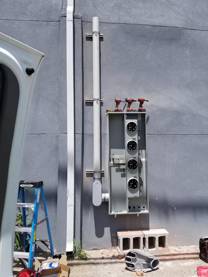 Electrical meter panel mounted on a gray wall with conduit and a ladder present.