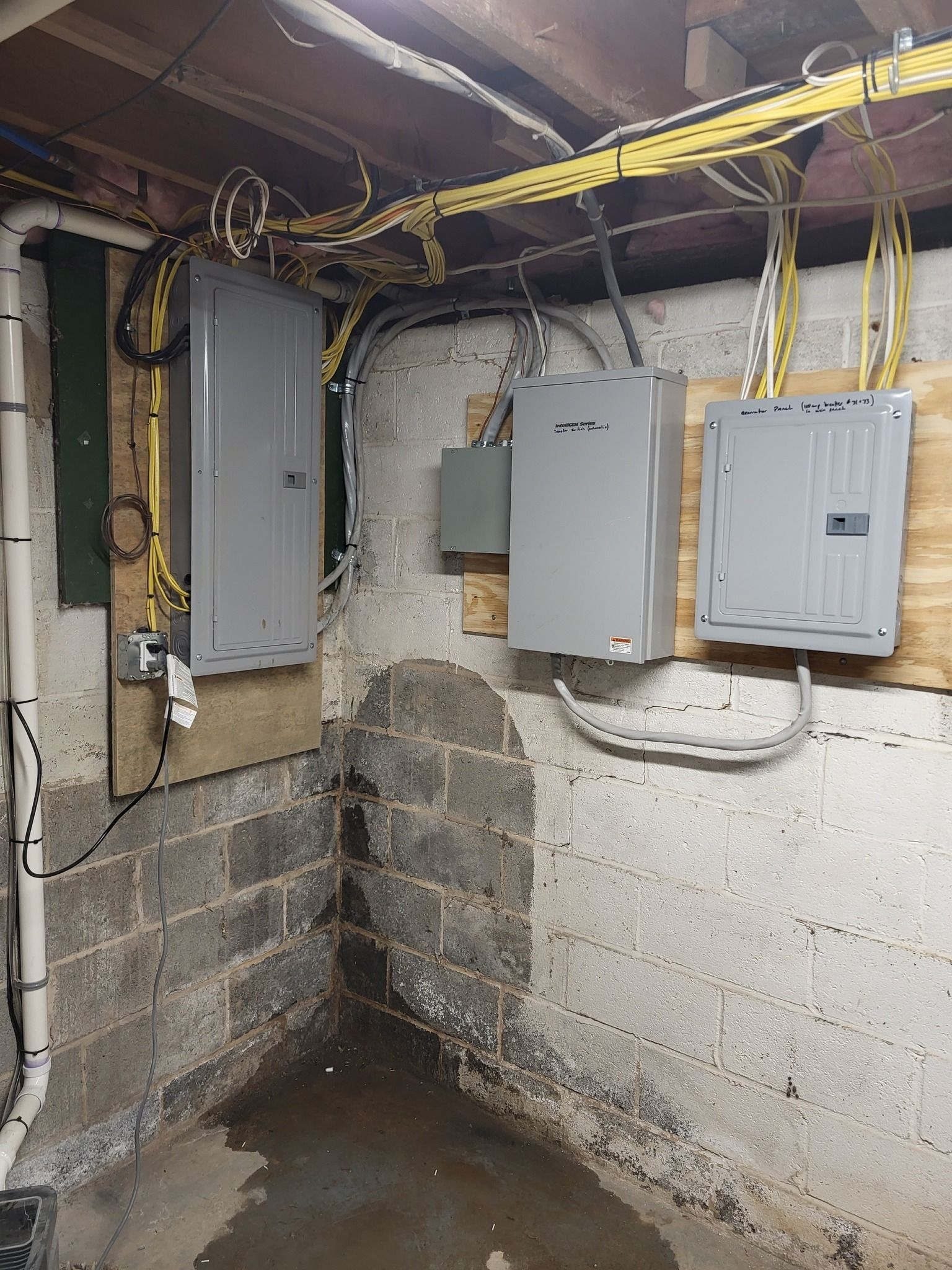 Electrical panels and wiring in a damp basement with concrete block walls and a wet floor.