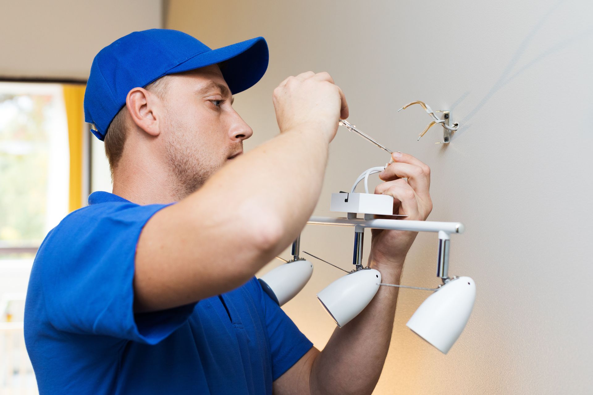 Electrician installing a ceiling light fixture with a screwdriver. White wall, blue shirt and cap.