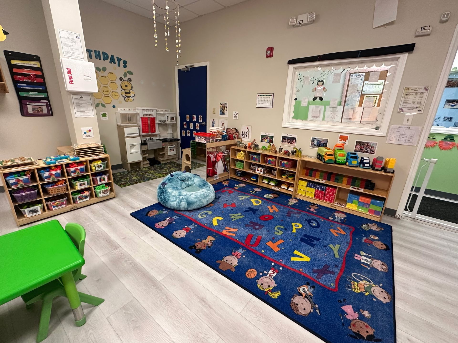 A brightly lit classroom with a blue alphabet rug, toy shelves, a green table, and a play kitchen area.