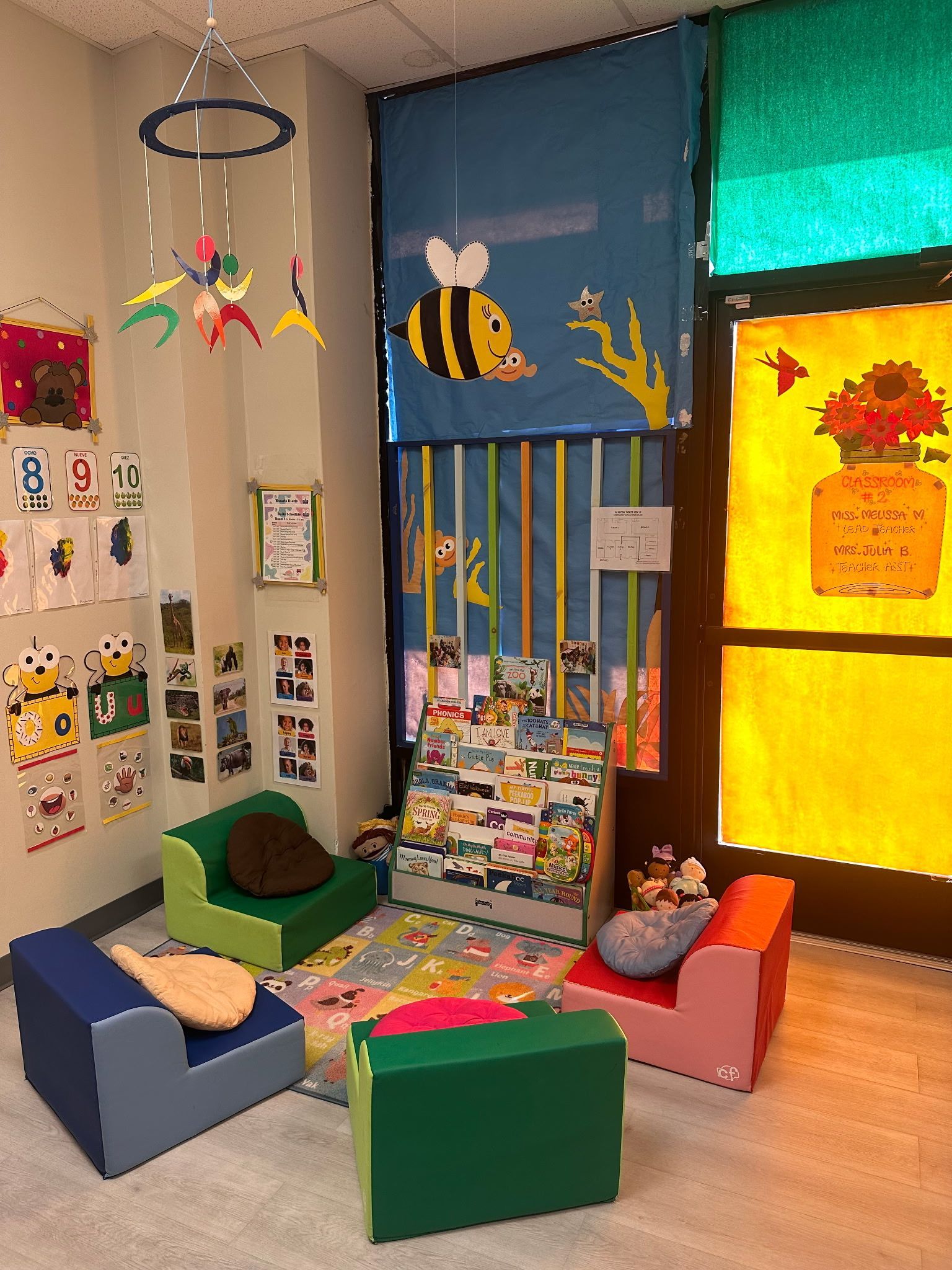 A small, colorful reading nook for children featuring soft chairs, a book display, and decorations of a bee and flowers.