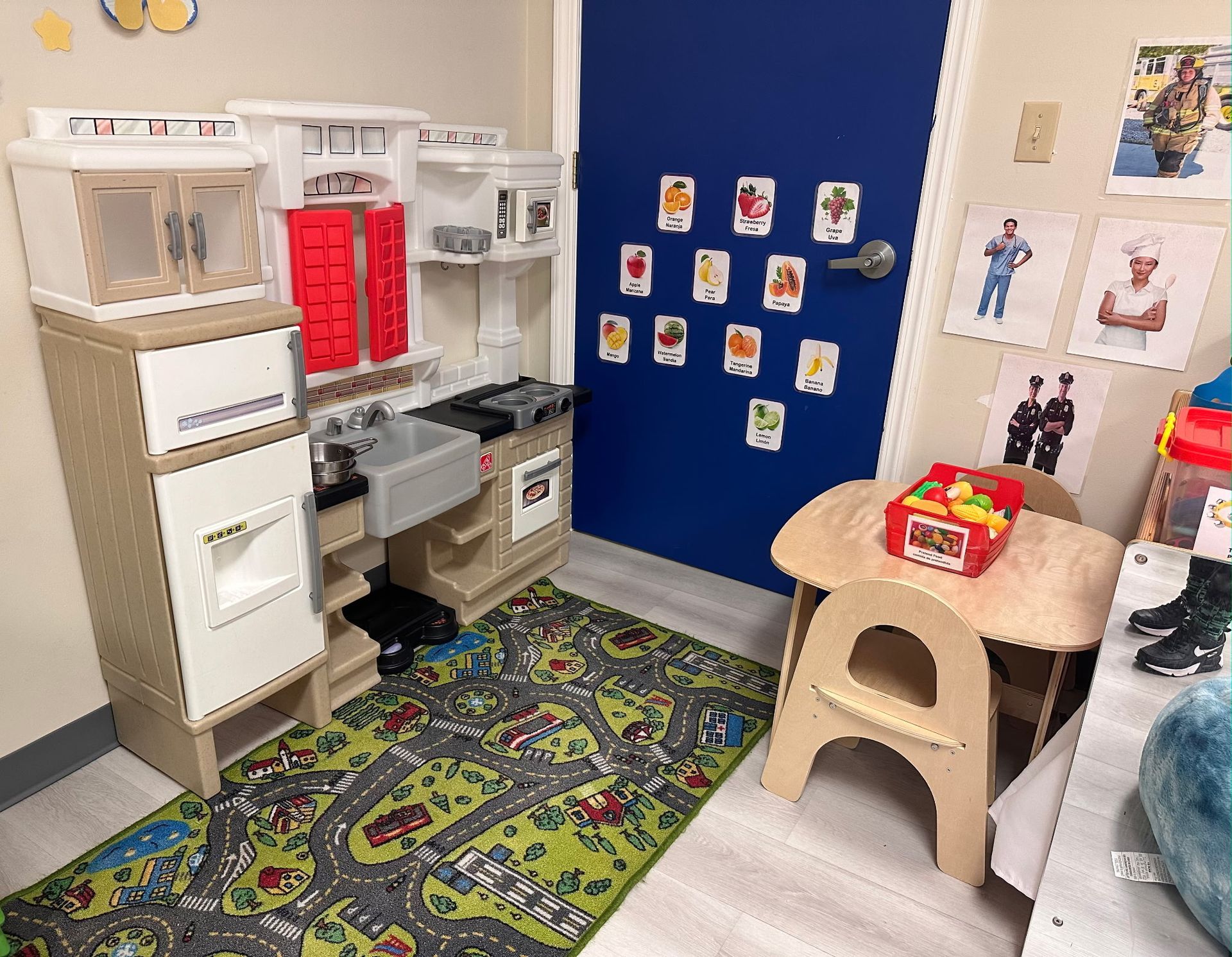A play kitchen and child-sized table on a rug with a road print, placed in front of a blue door decorated with pictures.