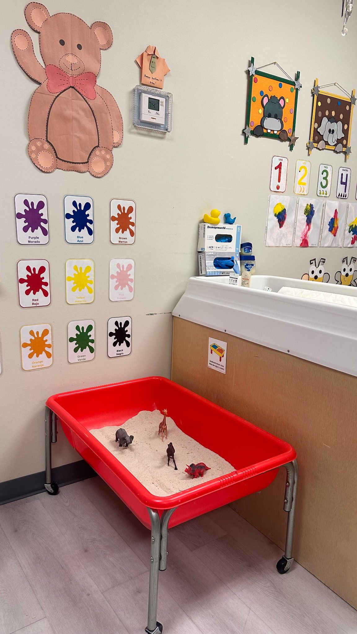 A red sensory table with sand and toy animals in a classroom with educational wall decorations and a changing station.