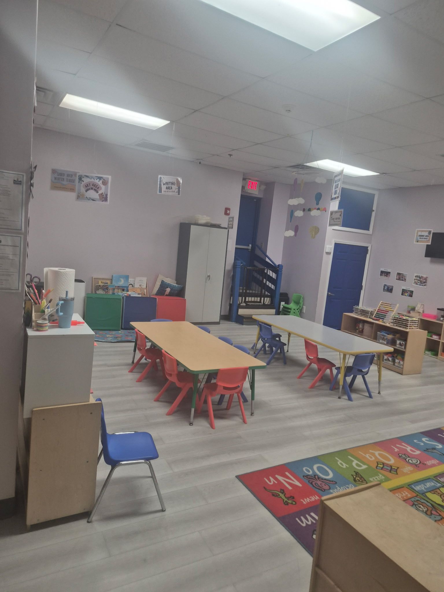 An indoor preschool classroom with tables, chairs, shelving units, and a colorful alphabet rug on the floor.