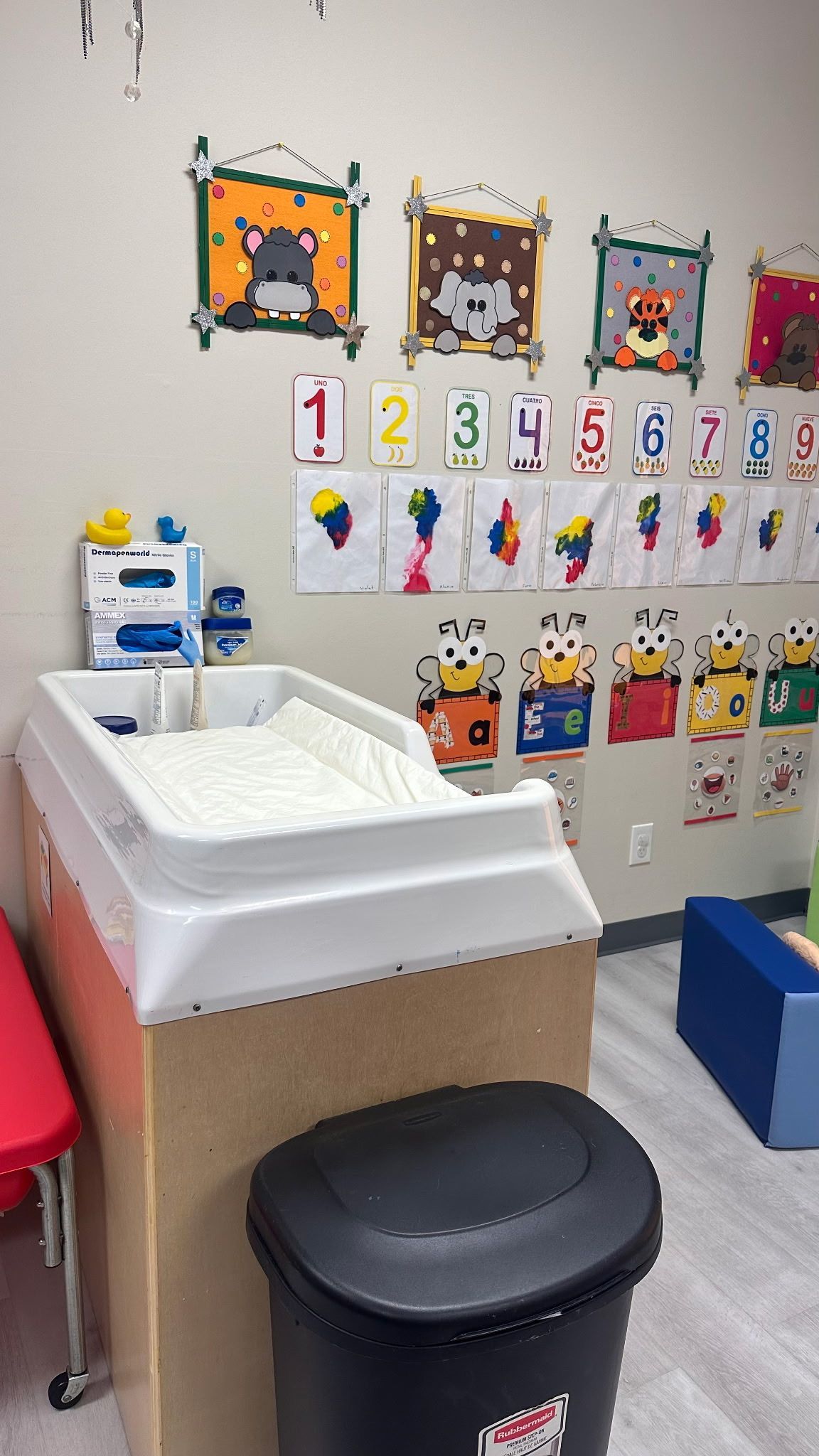 A white changing table stands beside a black waste bin in a child's classroom decorated with colorful animal art.