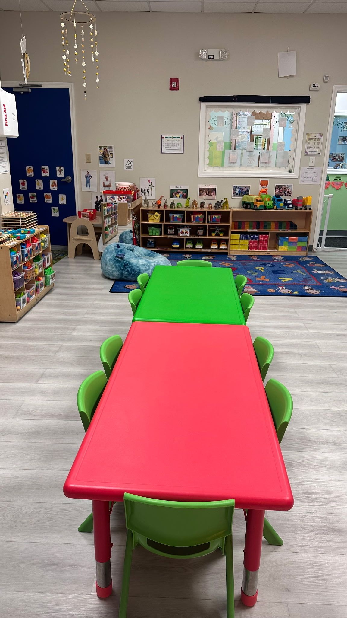 A preschool classroom featuring a long red table and a smaller green table with matching green chairs on a light floor.