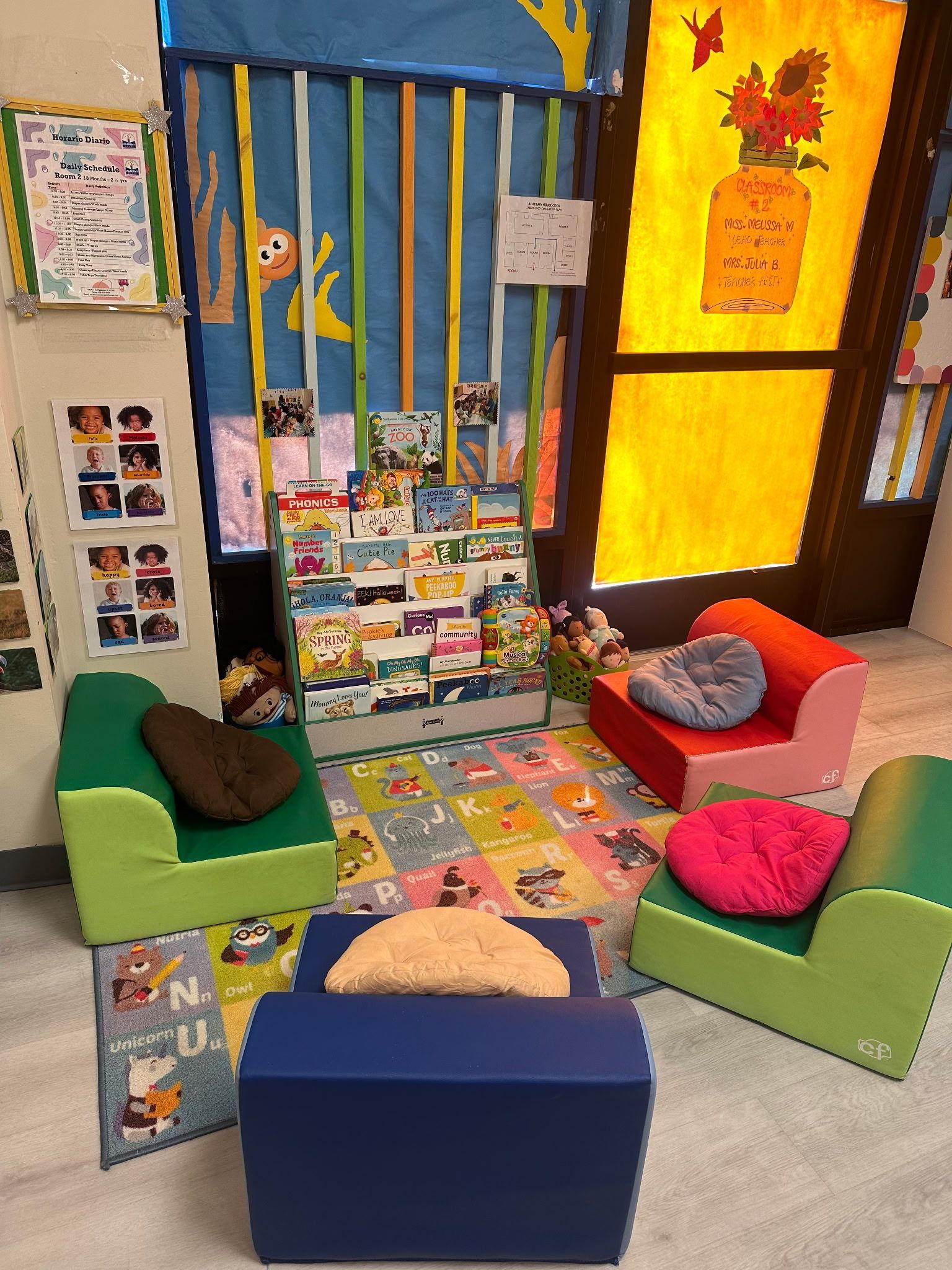 A reading corner with four colorful chairs, a book rack, and a patterned rug in a bright classroom setting.