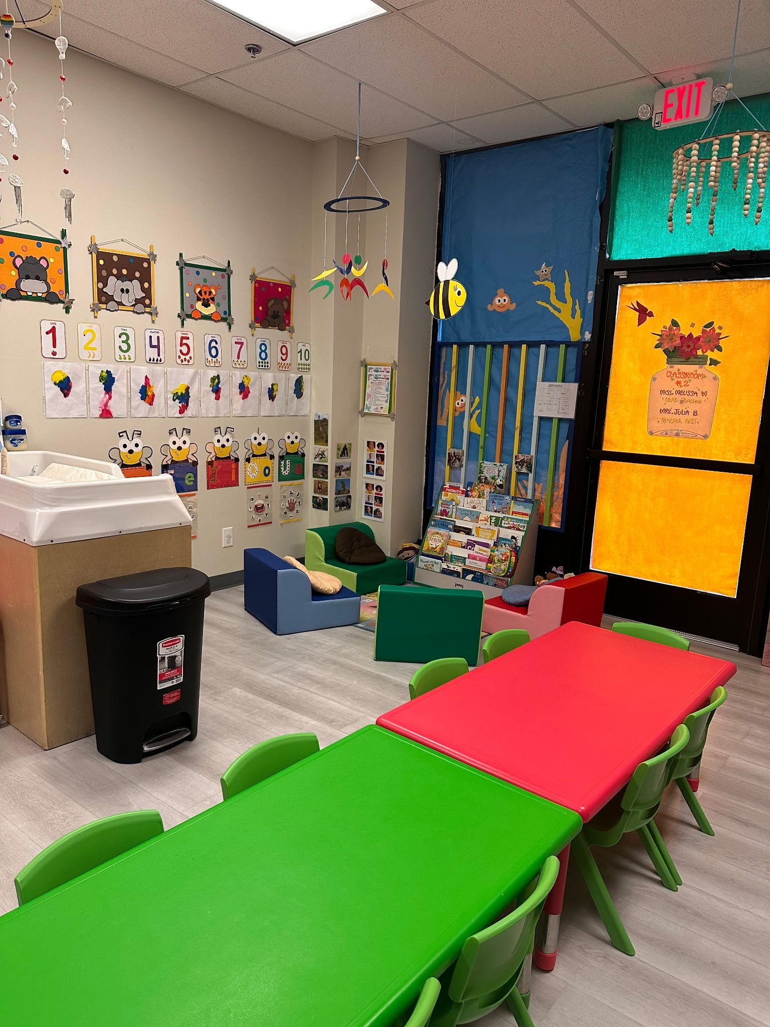 A colorful classroom with green and red tables, small chairs, and wall-mounted art displays.
