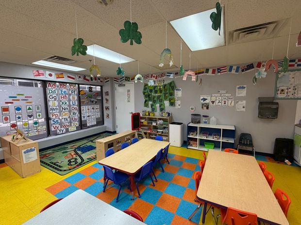 A brightly lit classroom with two tables, colorful checkered flooring, shamrock decorations, and educational wall charts.