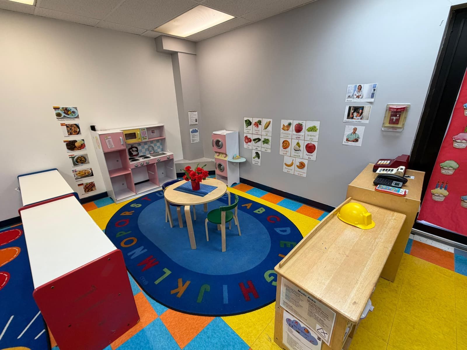 A brightly colored classroom corner with a circular rug, small table and chairs, toy stations, and wall posters.