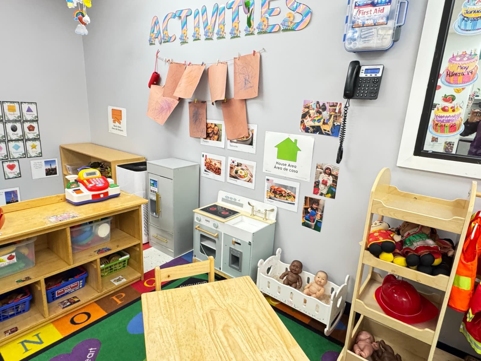 A colorful preschool play area featuring a toy kitchen, a toy cash register, a small table, a doll bed, and craft displays.