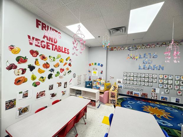 A colorful preschool classroom with "Fruits and Vegetables" wall art, student desks, and a sun-themed area rug.