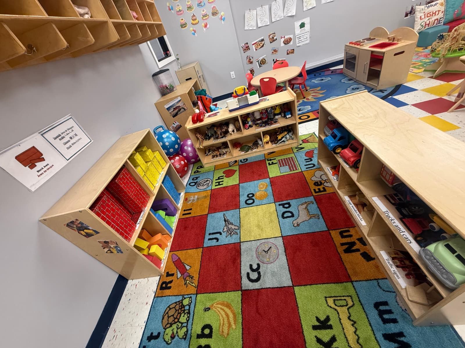 A colorful preschool classroom area features wooden shelves filled with toys, positioned on a large alphabet-patterned rug.