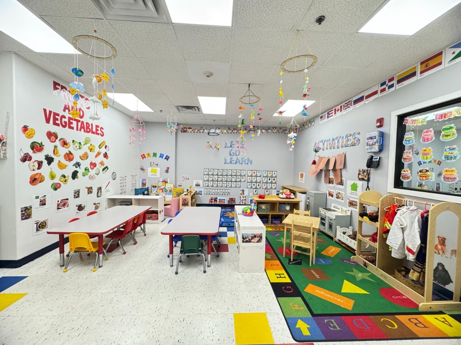 A colorful, brightly lit preschool classroom featuring tables, chairs, learning posters, and an educational area rug.