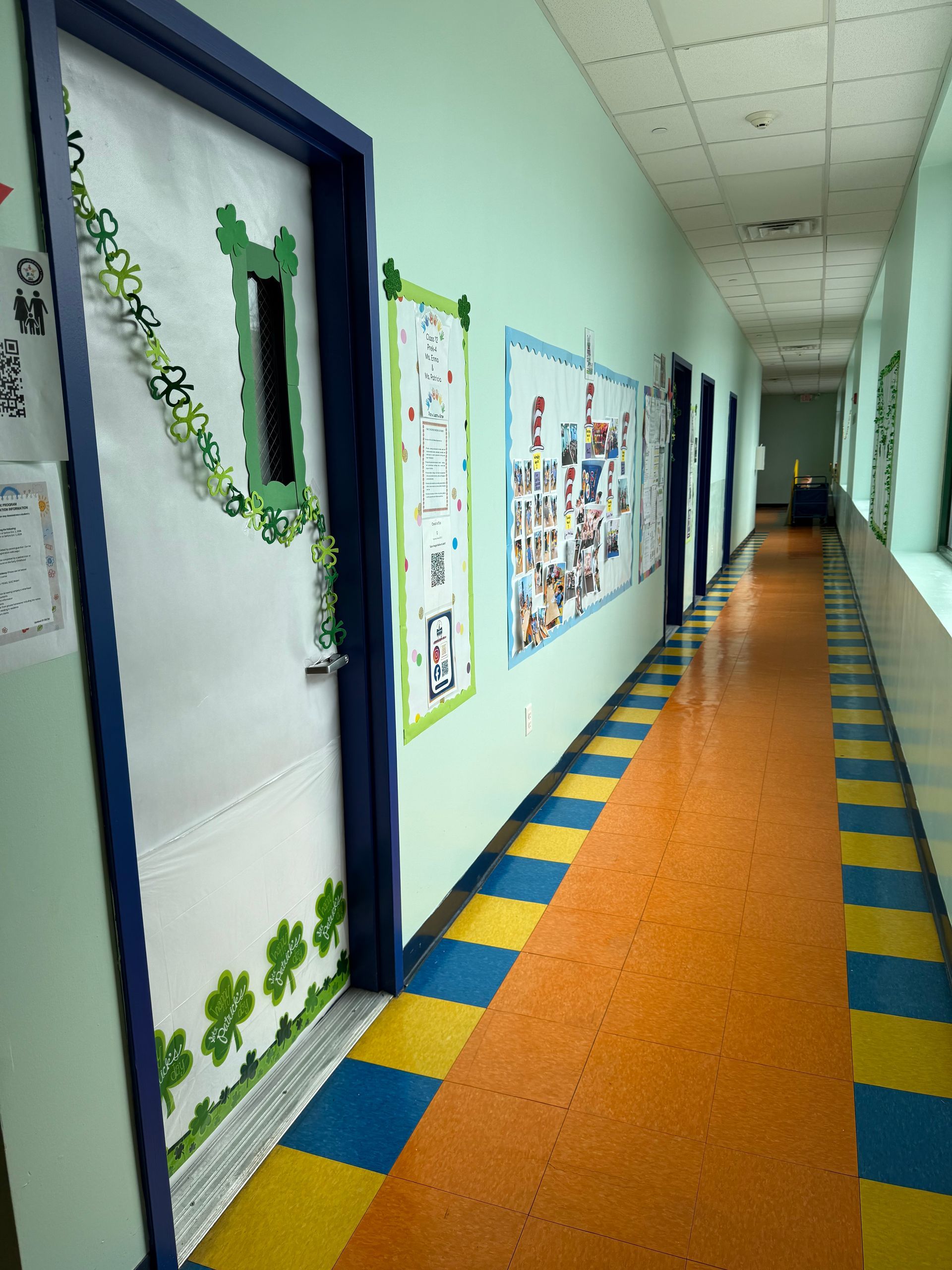 A brightly lit school hallway with blue and yellow checkered floors, pale green walls, and doors decorated for St. Patrick's.