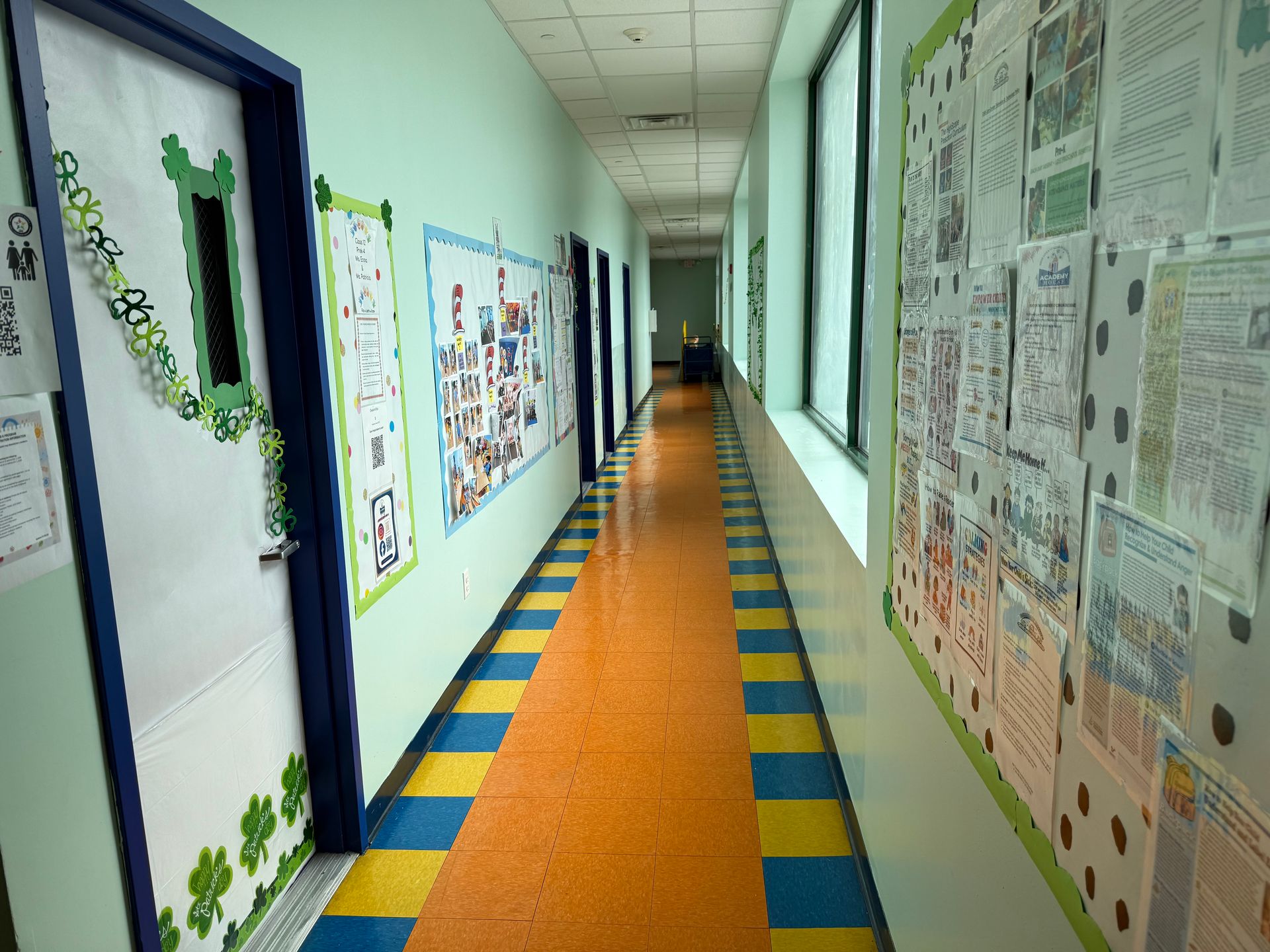 A brightly lit school hallway with a multi-colored tile floor, decorated doors, and walls lined with posters.