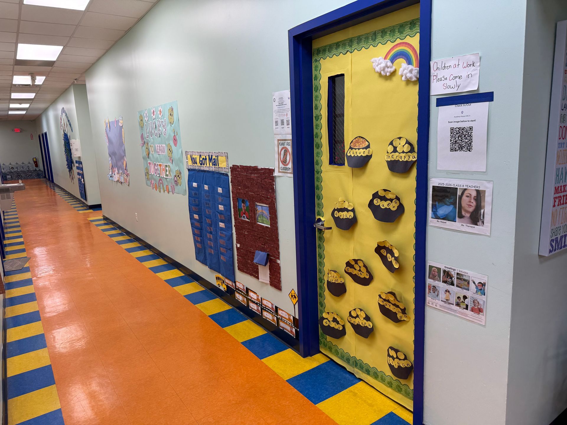 A brightly lit school hallway with orange and blue floors, featuring a yellow classroom door decorated with rainbows.