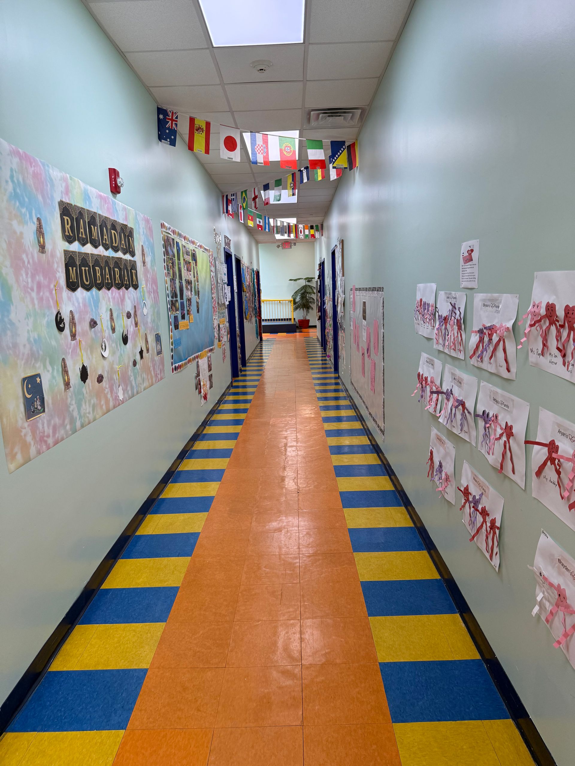 A school hallway with flags hanging from the ceiling, walls covered in children's art, and a blue and yellow striped floor.