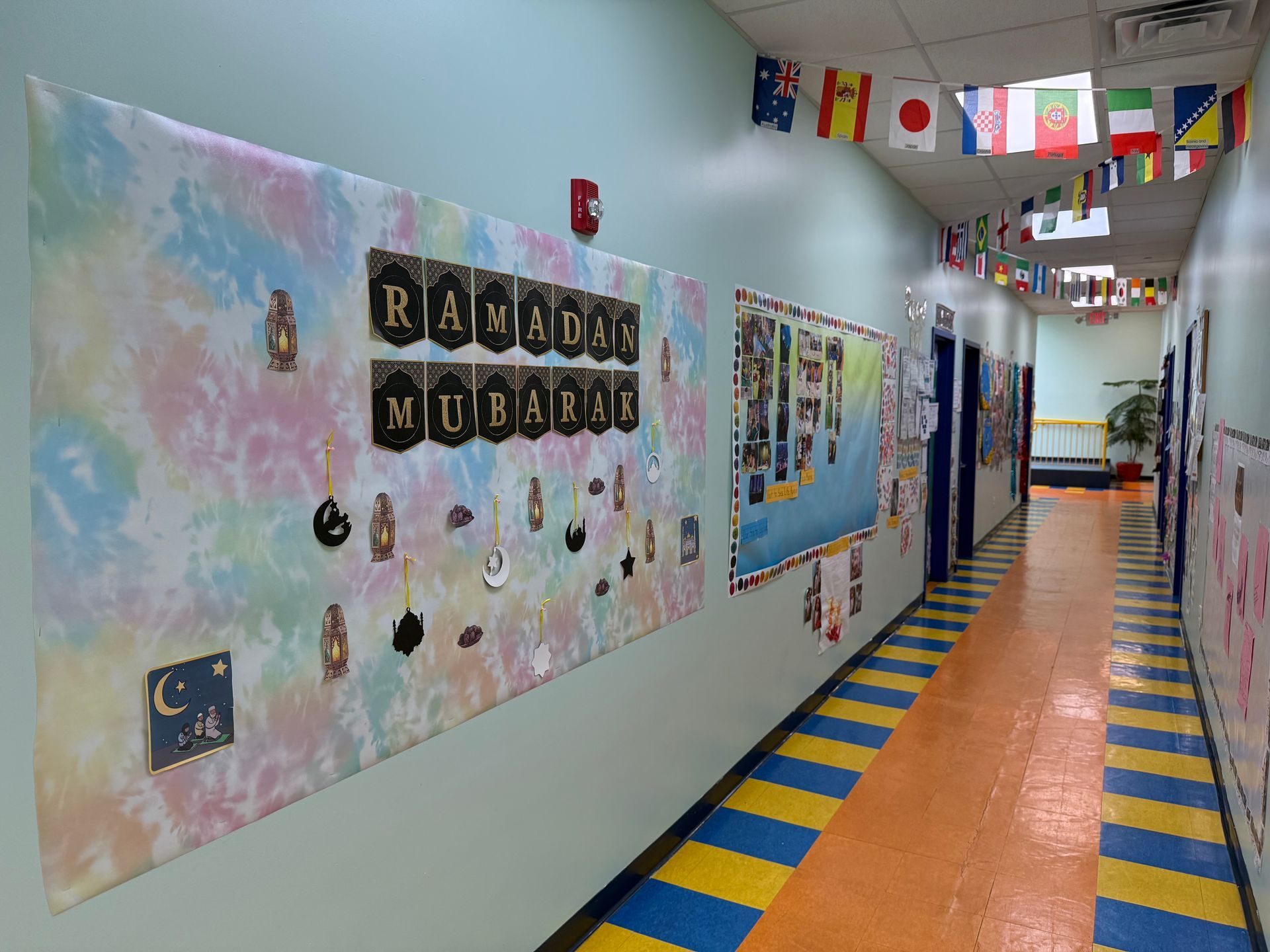 A school hallway decorated with a tie-dye Ramadan Mubarak bulletin board and international flag banners hanging above.
