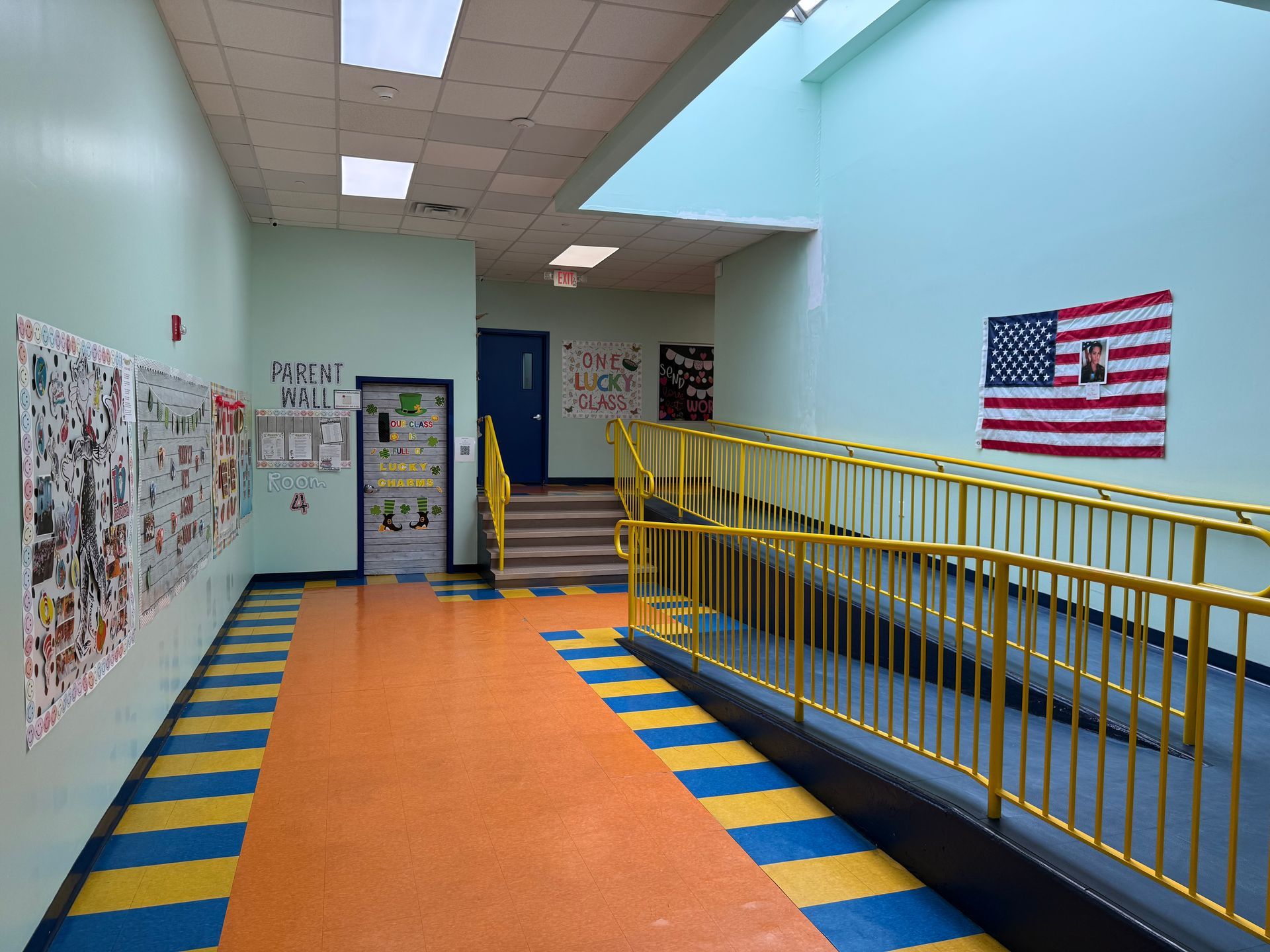 A school hallway with a yellow-railed ramp, blue-striped orange flooring, colorful wall posters, and an American flag.