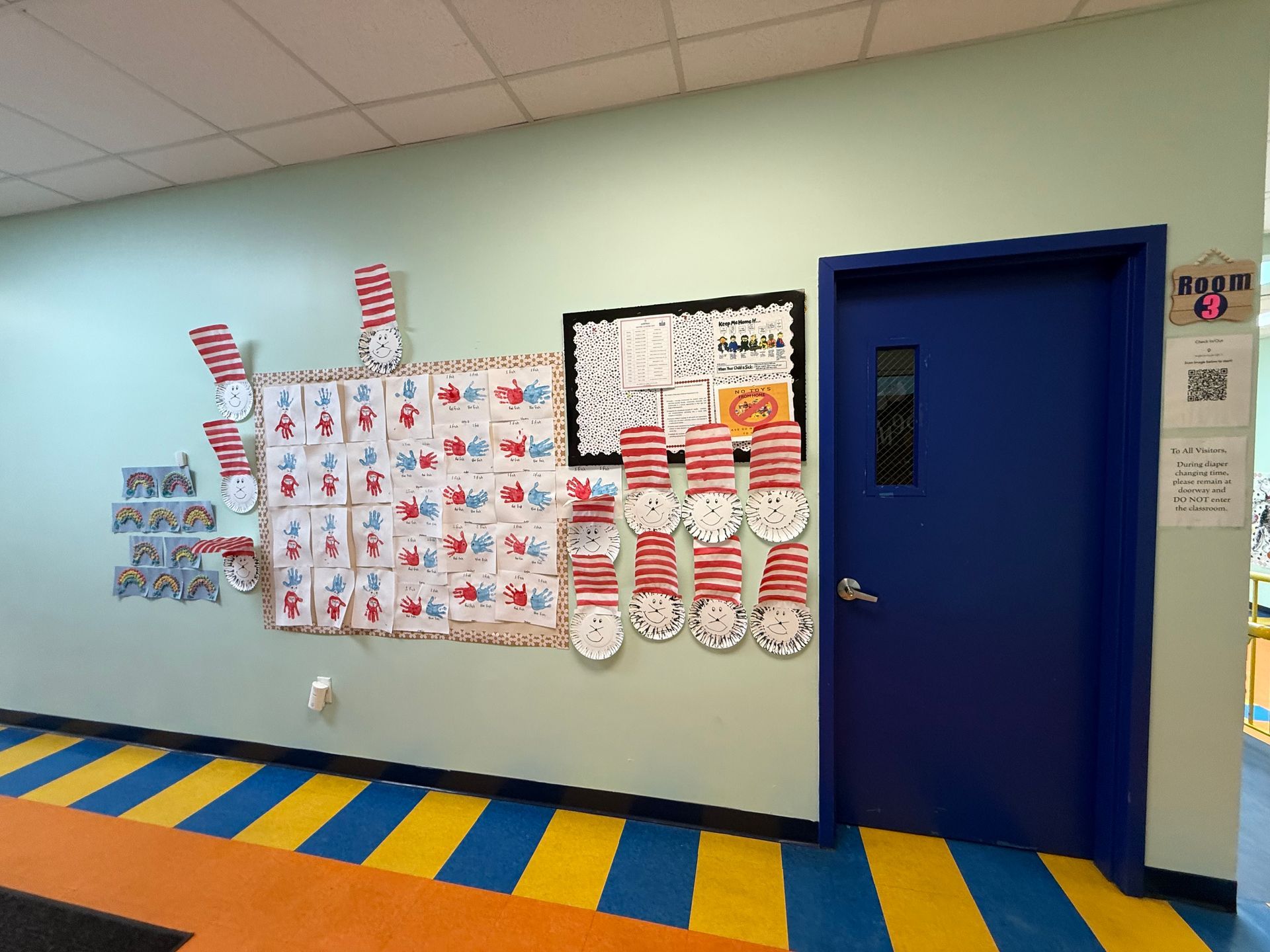 A decorated hallway wall features student art projects with red-and-white hats next to a blue classroom door.