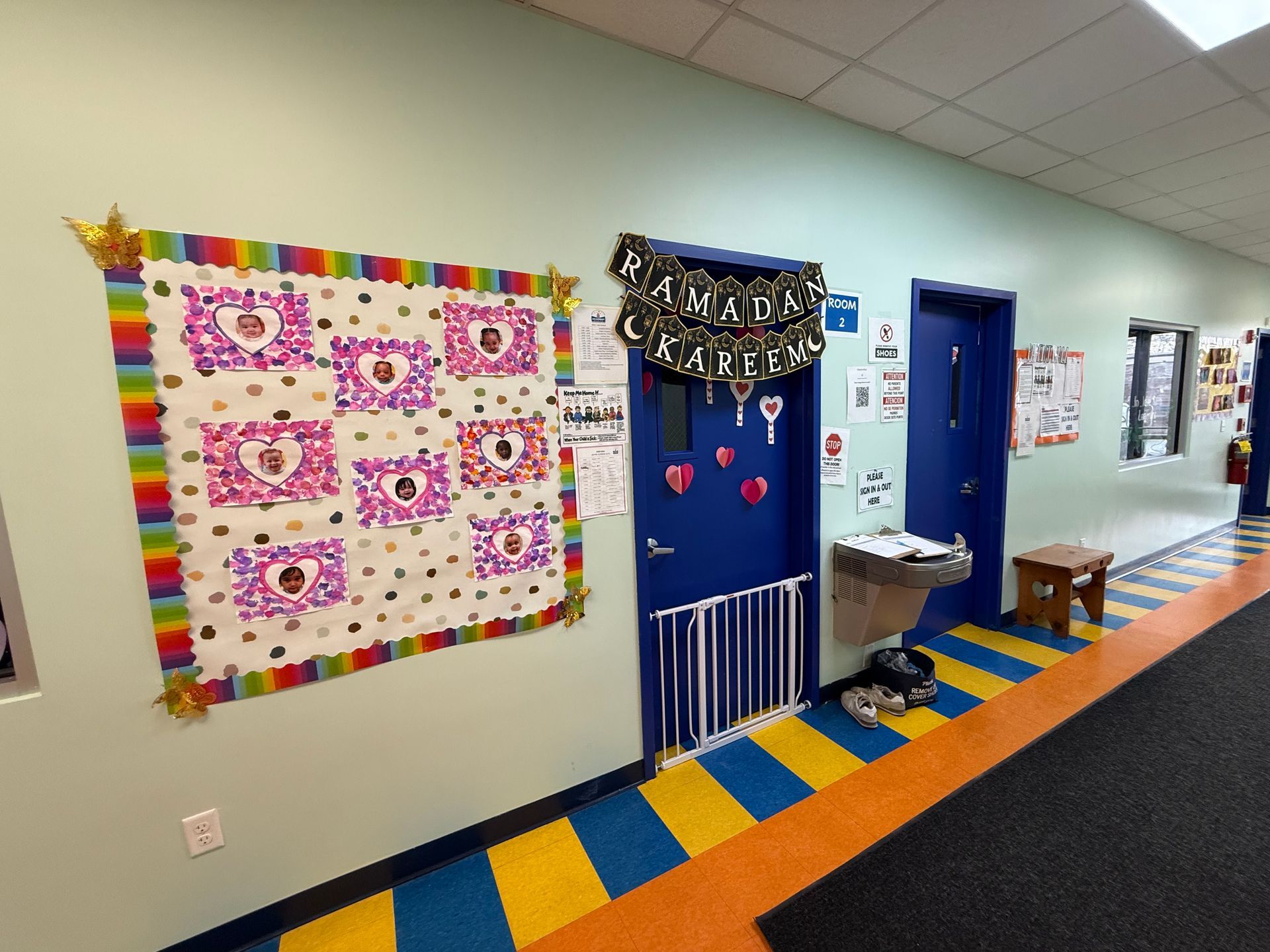 A school hallway with a rainbow-bordered bulletin board, a blue door decorated for Ramadan, and a small wooden stool.