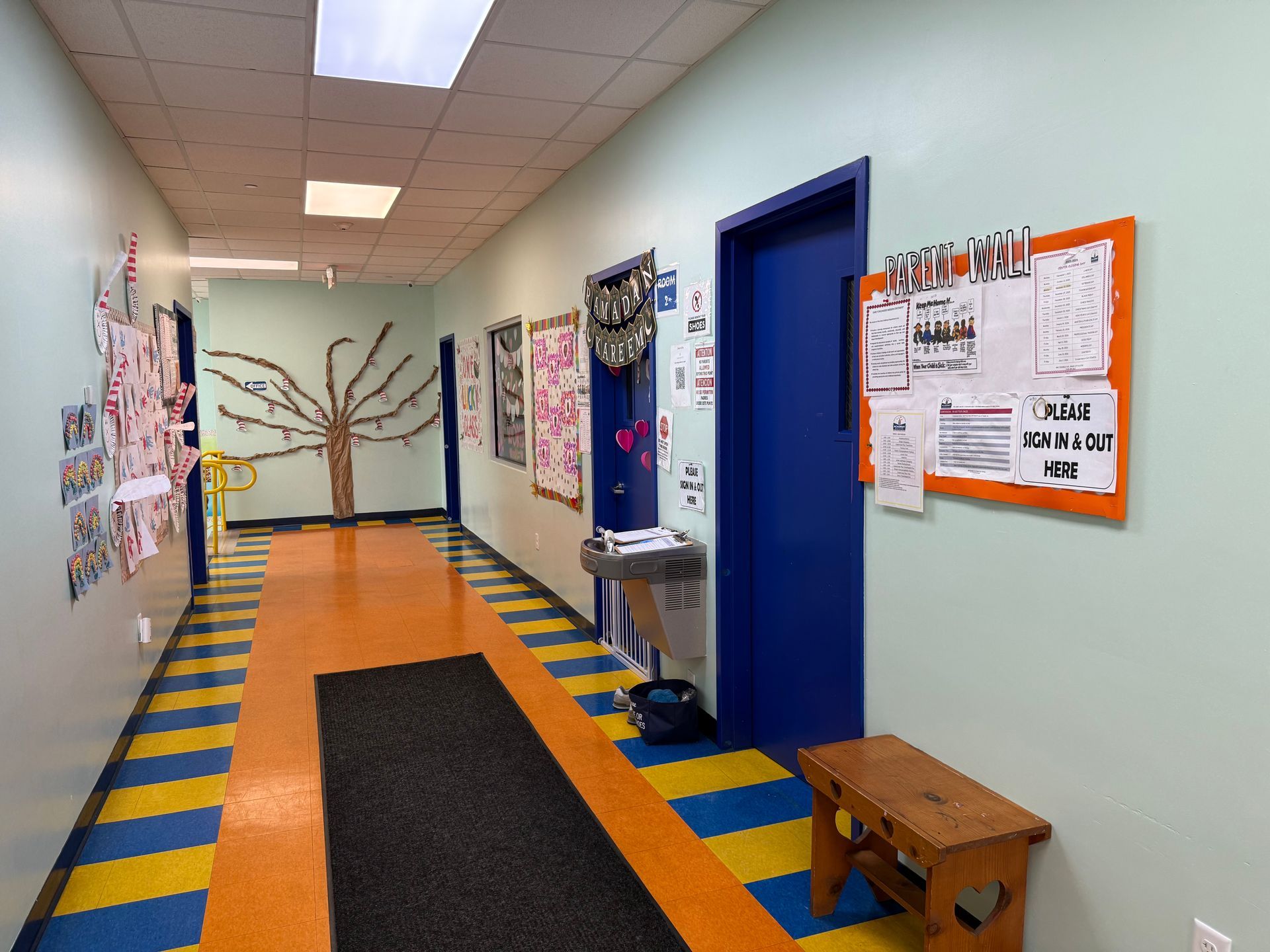 A brightly lit school hallway with blue and yellow striped floors, a decorative tree on the wall, and an orange bulletin.