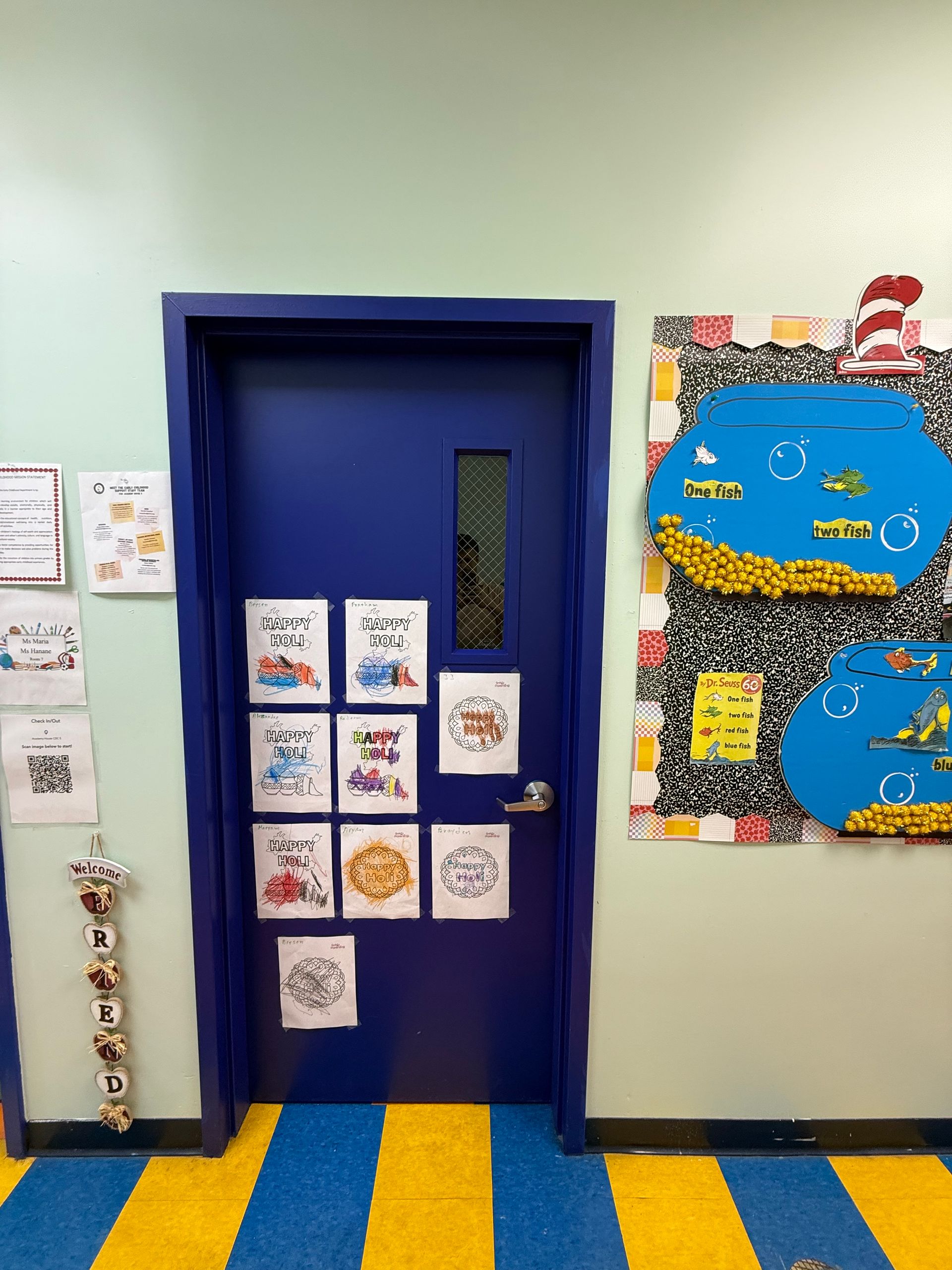 A blue classroom door decorated with student artwork and a Dr. Seuss bulletin board on a yellow and blue striped floor.