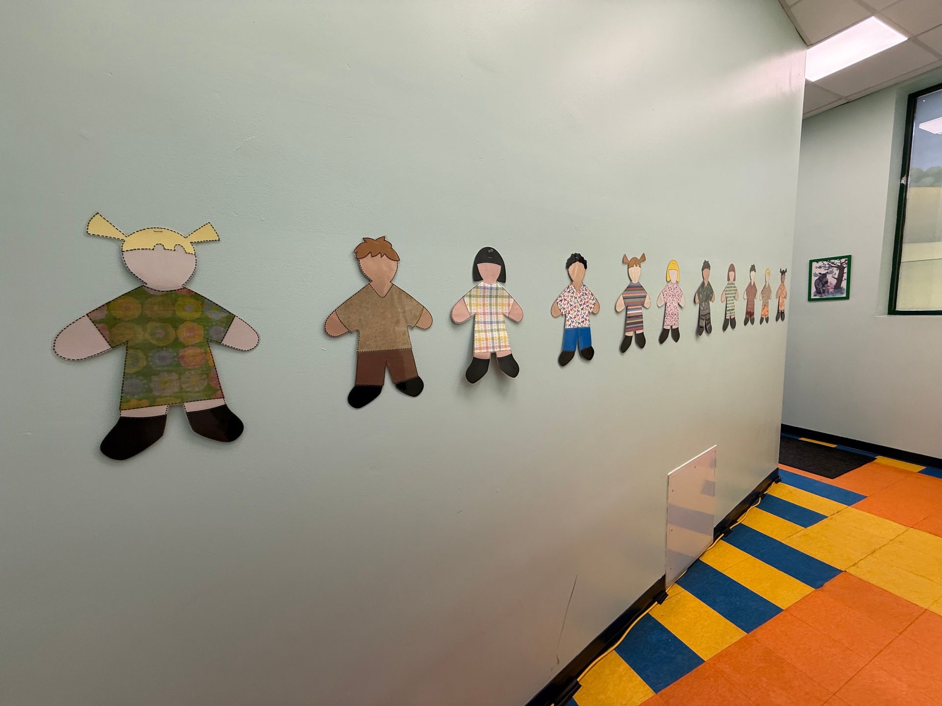 A row of paper cutout figures of various sizes and patterned clothing displayed on a light blue wall.