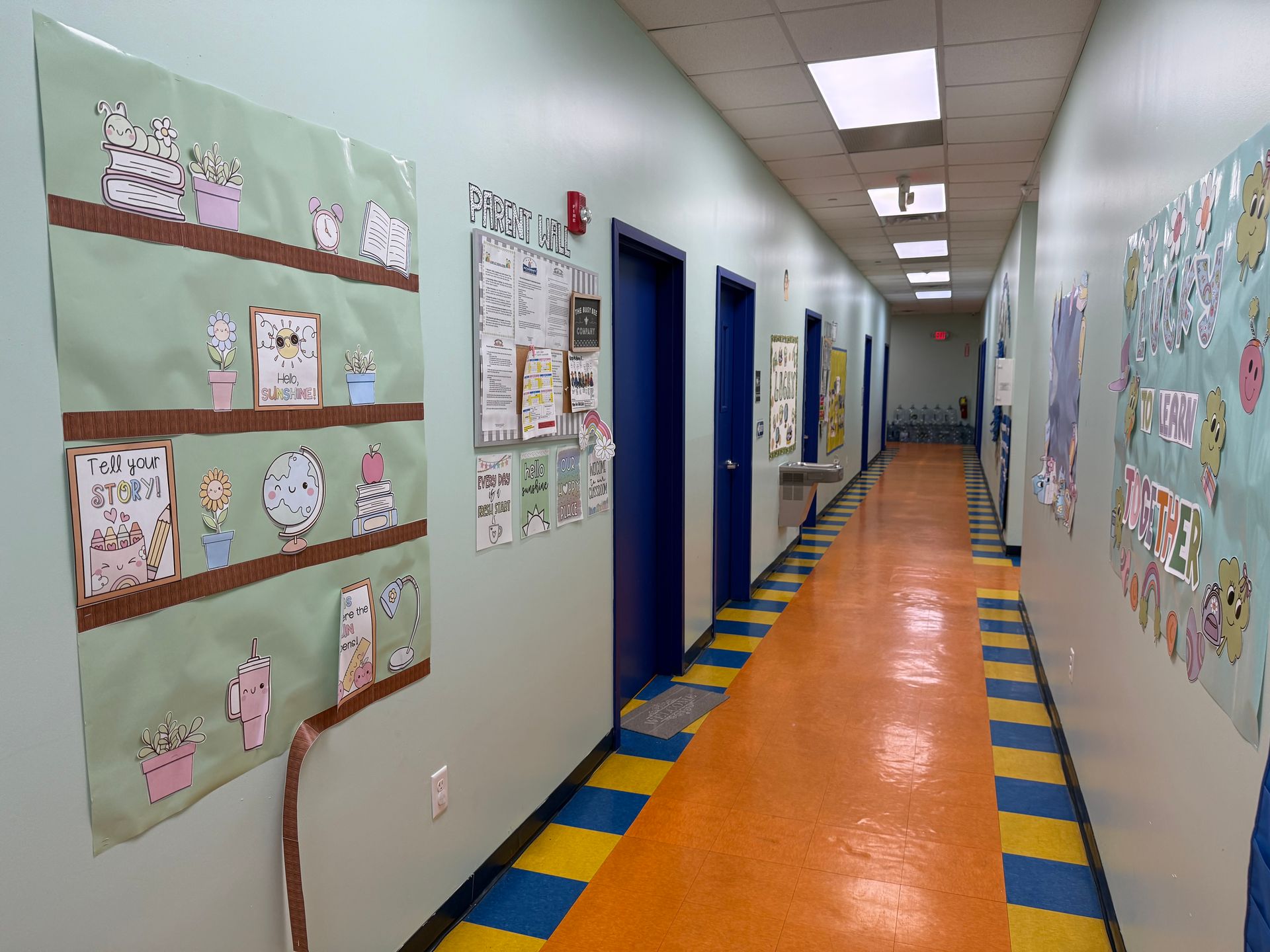 A school hallway with light green walls, a bright orange floor, blue doors, and colorful student artwork on the walls.
