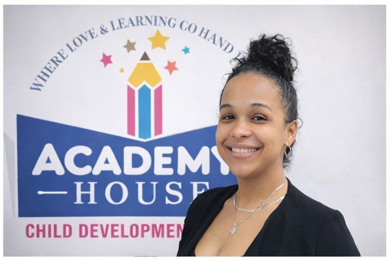 A smiling person stands in front of an Academy House Child Development logo with a colorful pencil and star design.