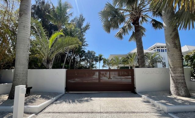 A brown metal gate on a white wall, leading to a white house, flanked by palm trees.