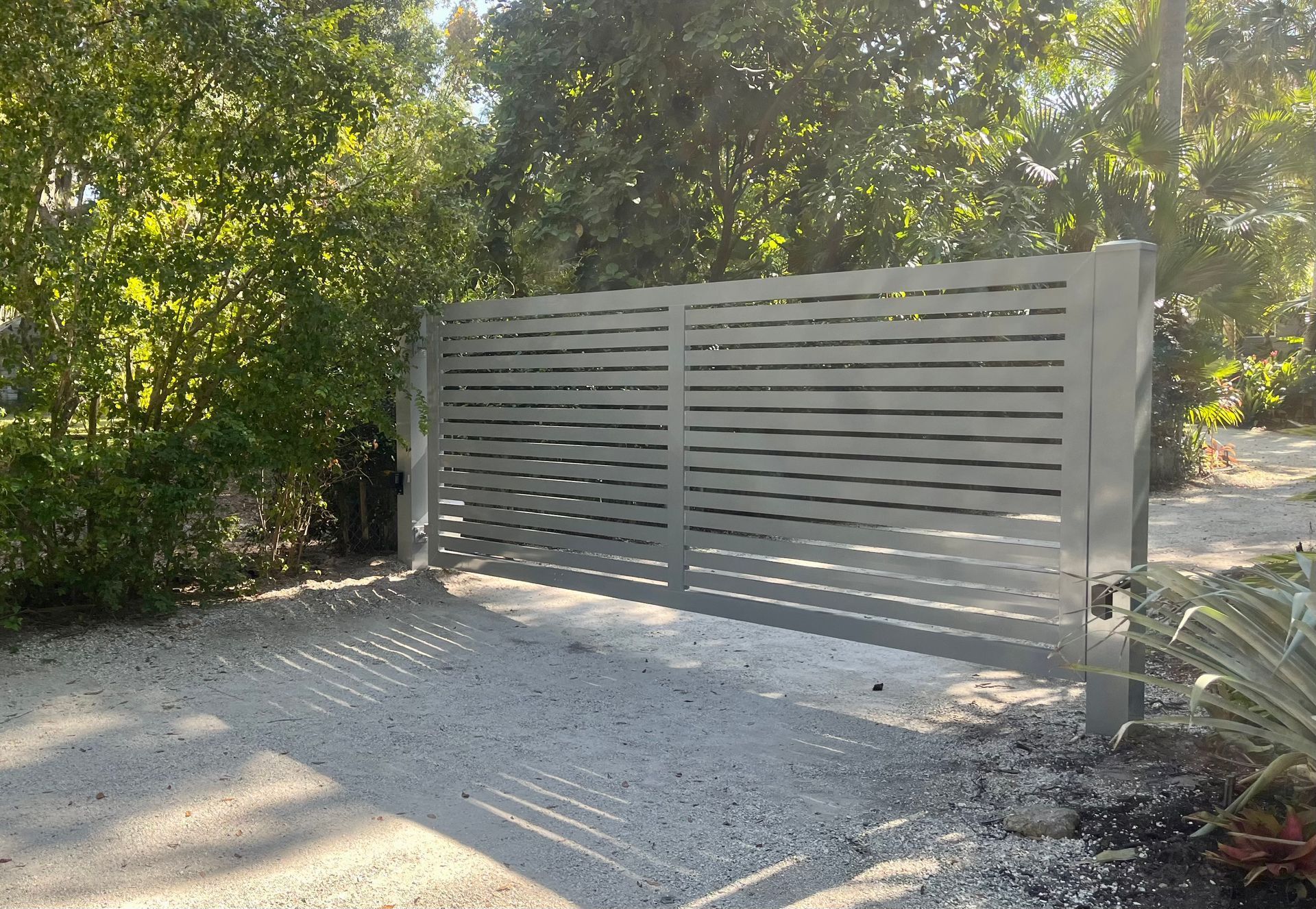 Gray horizontal slat gate, on a gravel driveway, set against lush green foliage.