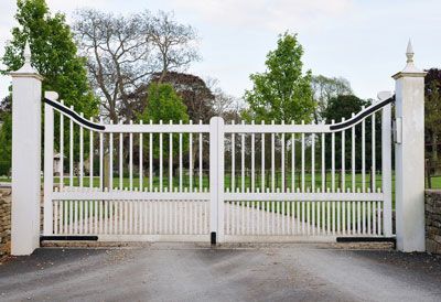 A white gate is open to a driveway with trees in the background