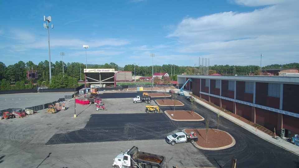 Construction site with trucks, new asphalt, and building. Bleachers and scoreboard in background.